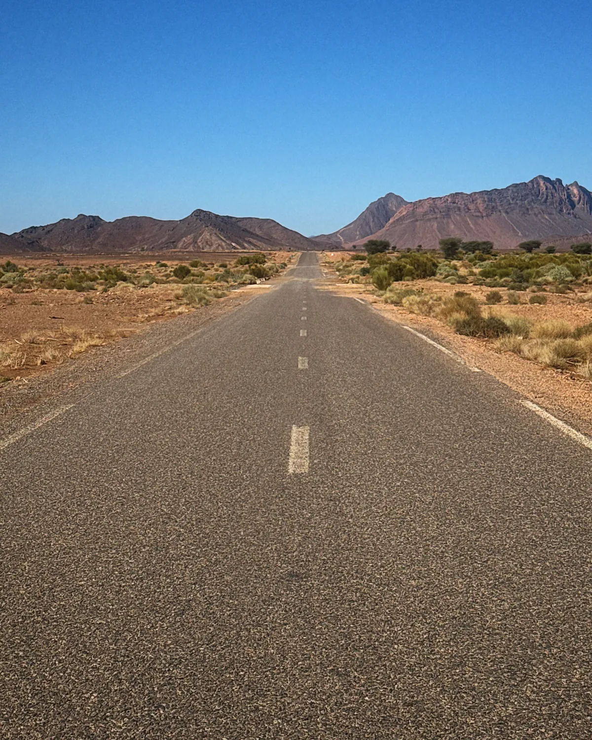An empty straight road across the Moroccan desert towards red mountains under a deep blue sky