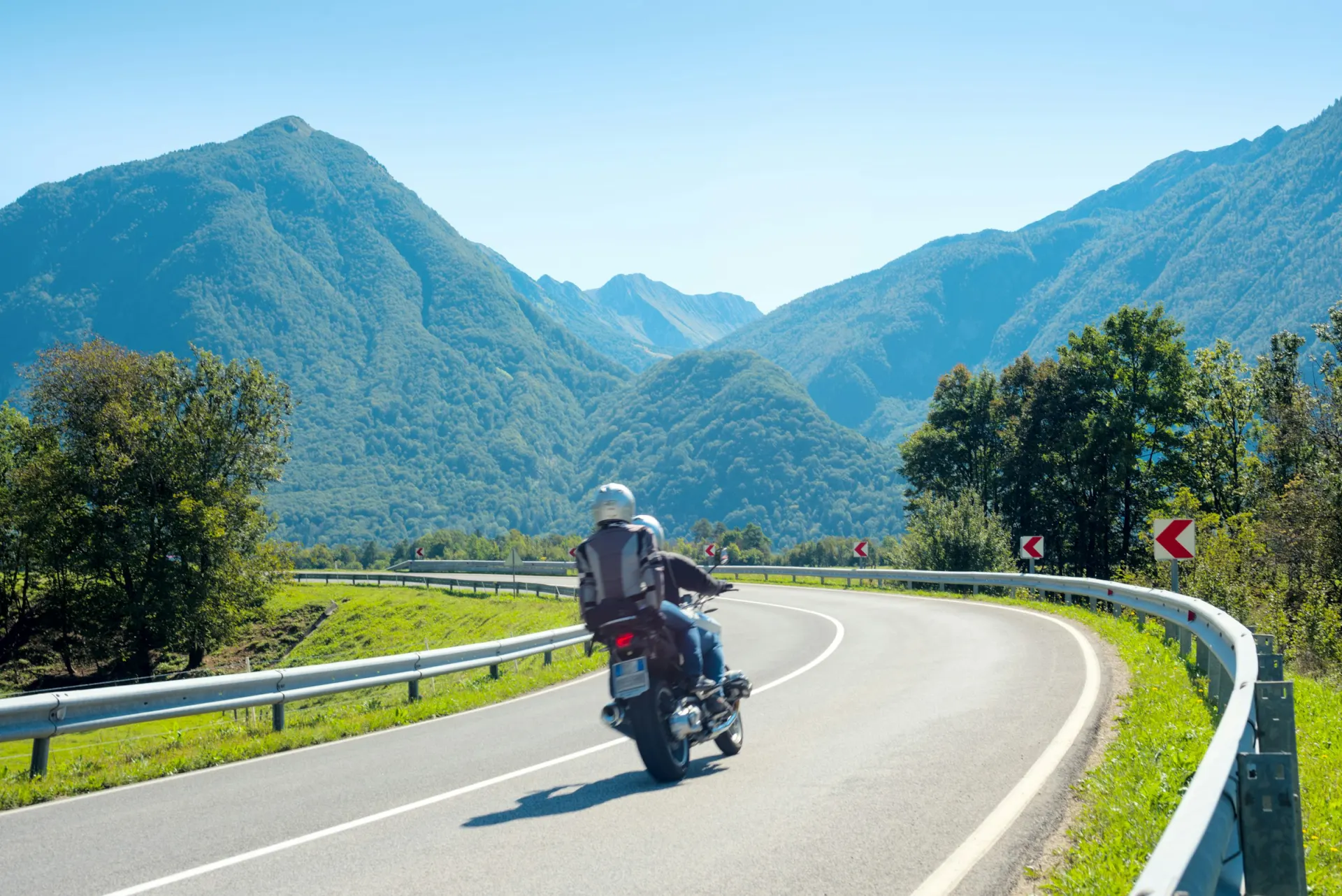 A rider and passenger on a motorcycle taking a curve on a mountain road under a clear blue sky